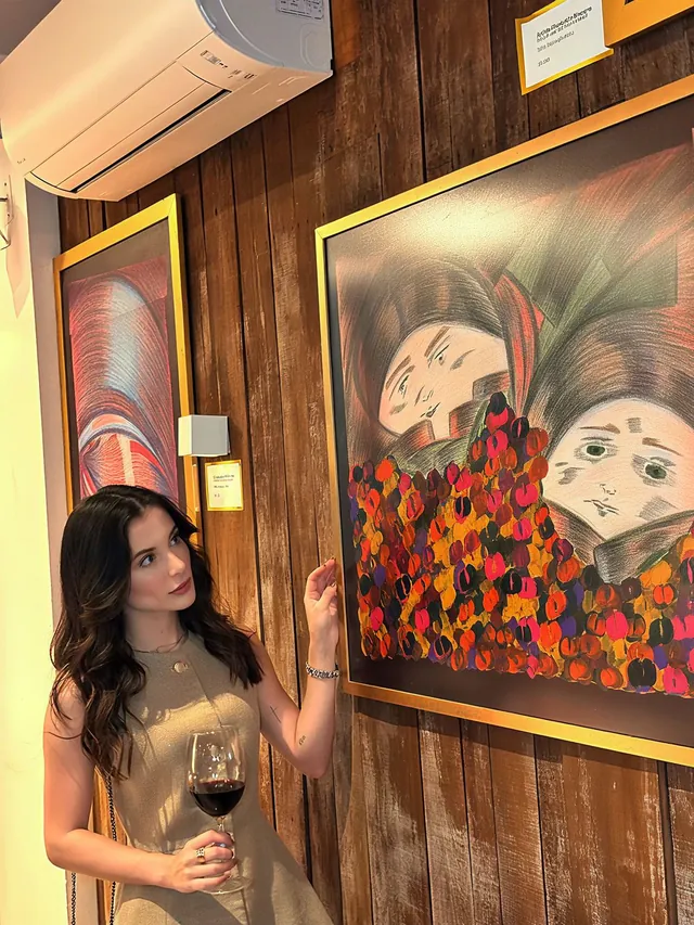 Bárbara Maria Vieira ao lado de pinturas em uma exposição de artes visuais