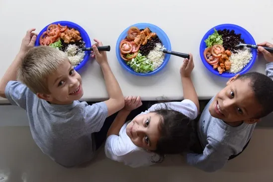 Vista de cima de três crianças sorrindo ao redor de pratos de refeição em uma mesa